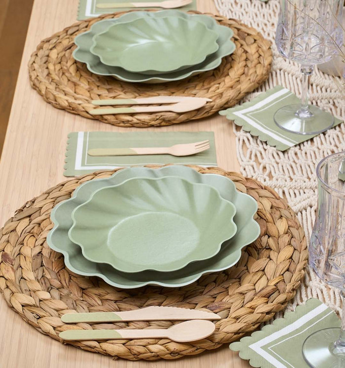 A table set with Sophistiplate's Cocktail Napkin Scalloped Edge Sage, woven placemats, green scalloped plates, eco-friendly wooden cutlery, compostable tableware, and clear glasses arranged in a light, natural style.