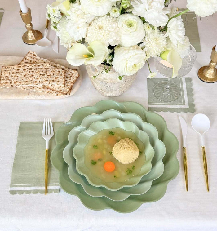 The Eco-friendly Table Setting- Sage by Sophistiplate LLC features pastel green scalloped bowls with matzo ball soup, gold flatware, matzo crackers, a green napkin, and eco-friendly disposable plates on a white floral tablescape.