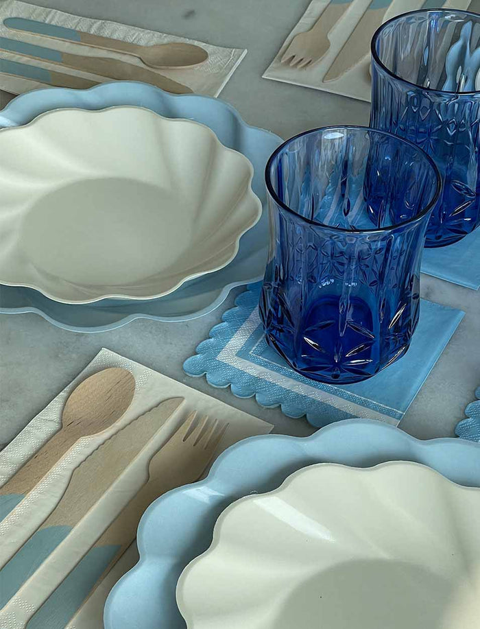 A table setting with white scalloped plates, blue chargers, eco-friendly tableware, blue glass tumblers, wooden cutlery, and Sophistiplate Cocktail Napkin Scalloped Edge Sky Blue arranged on a light surface.