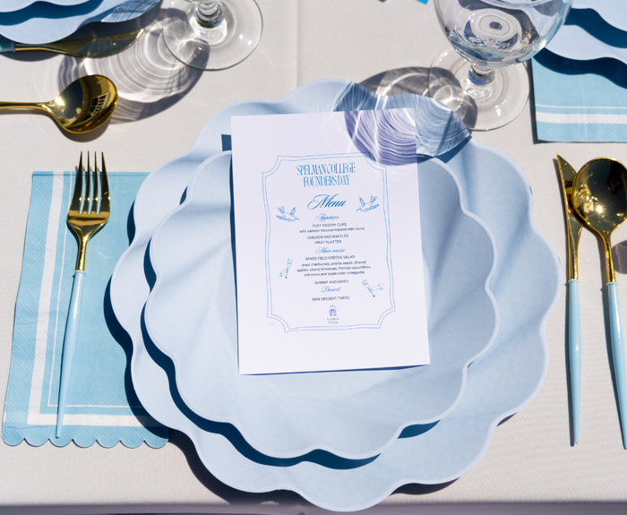 The Sophistiplate Eco-Friendly Table Setting- Sky Blue features plates with a printed menu, placed on a white tablecloth with gold cutlery, blue napkins, and clear wine glasses arranged neatly around it.