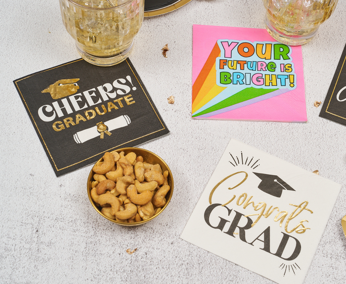 The Sugarplum Congrats Grad Cocktail Napkins (26pk) feature festive designs like “CHEERS! GRADUATE,” “YOUR FUTURE IS BRIGHT!” with a rainbow, and “Congrats GRAD.” Goldfish crackers and drinks are placed nearby.