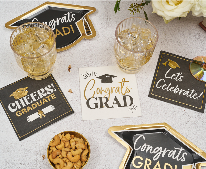 A tabletop decorated for graduation with “Congrats Grad” plates, Sugarplum Congrats Grad Cocktail Napkins (26pk), two iced drinks, a small bowl of cashews, and white flowers in the corner.
