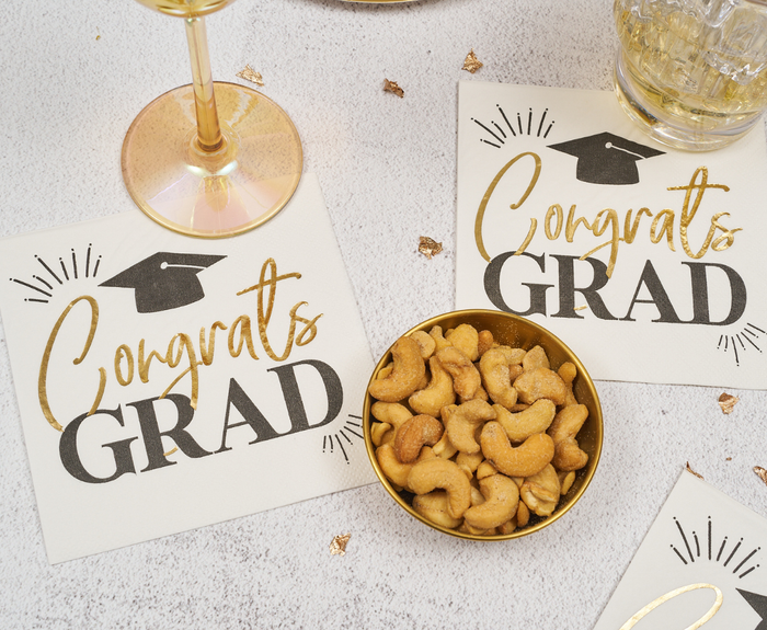 Sugarplum Congrats Grad Cocktail Napkins (26pk) in gold and black with a graduation cap design, shown on a light surface with gold confetti, a glass of drink, and a bowl of cashews.
