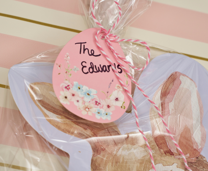 A Sugarplum Cookie Tray Gifting Kit Charming Easter/6pkg is wrapped in clear plastic with bunny ears and tied with pink and white string to a pink, floral Easter egg-shaped tag reading "The Edwards", set against a pink and white striped background.