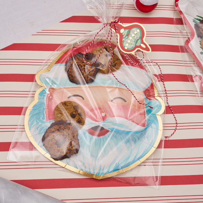 Sugarplum’s Cookie Tray Gifting Kit Santa/6pkg features chocolate chip cookies in a clear gift bag, tied with festive string and tag, resting on a Santa face card—just right for holiday treat exchanges on striped gift wrap.