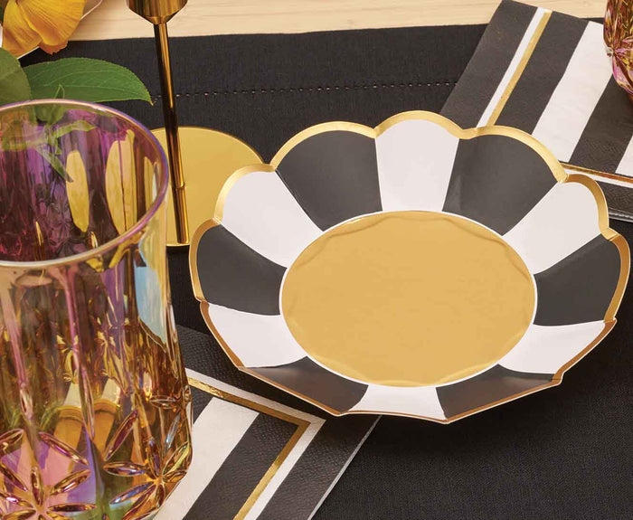 A Sophistiplate Cordoba Appetizer/Dessert Bowl from the 8pk set sits on a black and white striped napkin, next to an iridescent glass atop a dark tablecloth with a gold candle holder and green leaves.