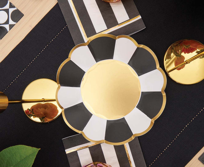 Sophistiplate’s Cordoba Appetizer/Dessert Bowls (8pk) feature a scalloped gold design, displayed on a dark, gold-edged textile with striped napkins, shiny gold accents, and a touch of green foliage at the edge.