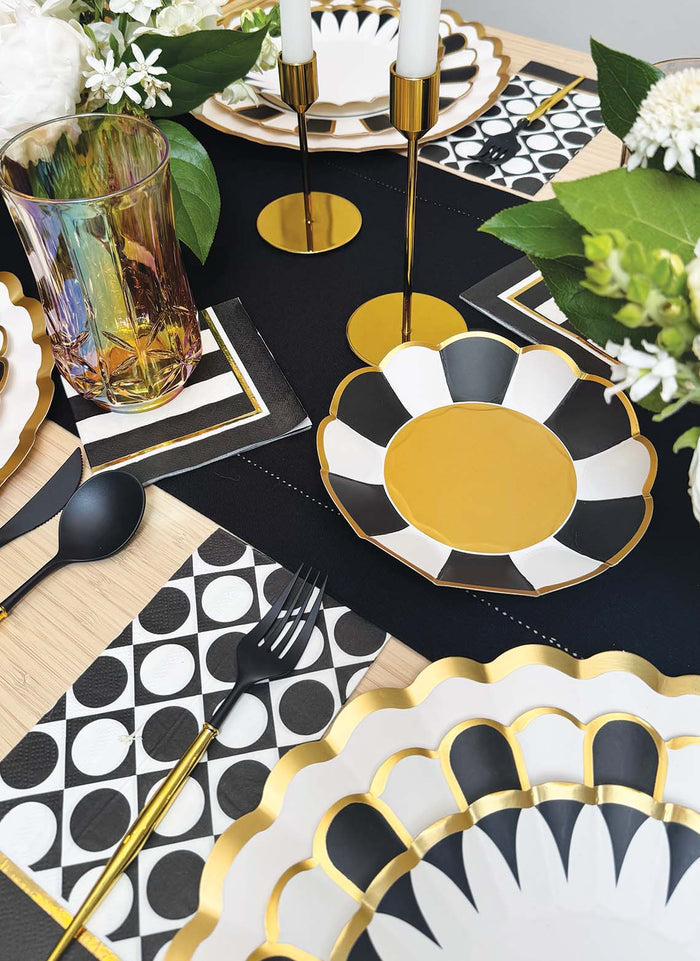 A stylish black and gold table features Sophistiplate Cordoba Appetizer/Dessert Bowls (8pk), flower-shaped plates, patterned napkins, black cutlery, iridescent glassware, gold candle holders, and elegant white floral decor.