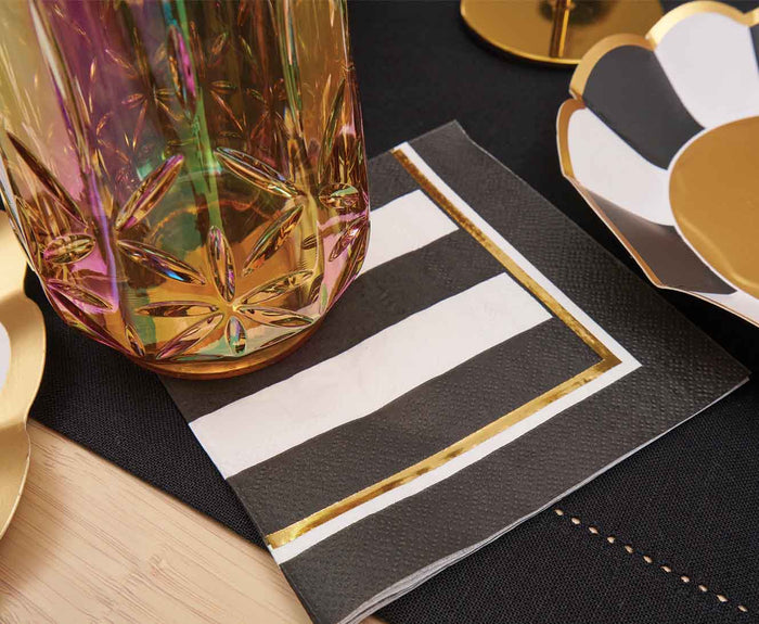 A close-up of a table set with Sophistiplate's Cordoba Cocktail Napkin/20pk, showcasing black-and-white stripes and a gold border, alongside an iridescent glass and a black and white plate with a gold center on a black tablecloth.