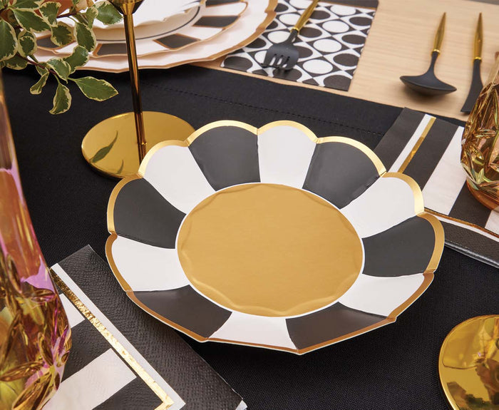 A scalloped plate with alternating black, white, and gold sections sits on a black placemat, surrounded by gold utensils, Sophistiplate Cordoba Cocktail Napkins, and gold candle holders atop a light wooden table.