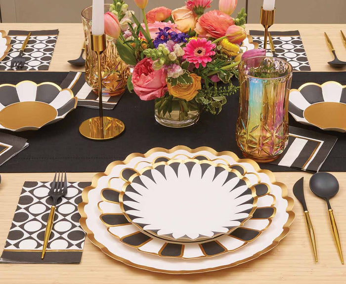 A modern table setting with Sophistiplate Cordoba Dinner Plates (8pk), gold flatware, black and white dotted napkins, colorful glasses, gold candle holders, and a vibrant floral centerpiece.