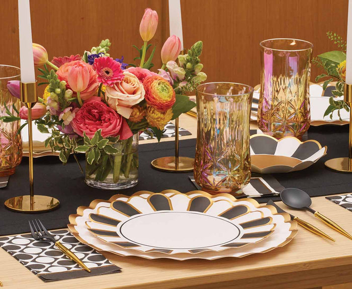 A table set with Cordoba Dinner Plates by Sophistiplate, iridescent tumblers, gold flatware, black placemats, black-and-white napkins, gold candlesticks with white candles, and a colorful flower arrangement.