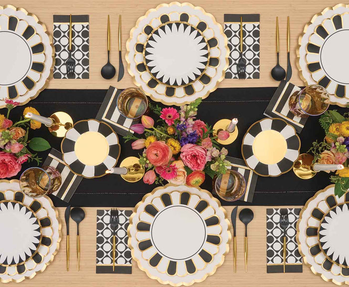 A stylish table setting featuring Sophistiplate Cordoba Dinner Plates, black cutlery, geometric napkins, gold-rimmed glasses, and a colorful floral centerpiece on a black table runner.