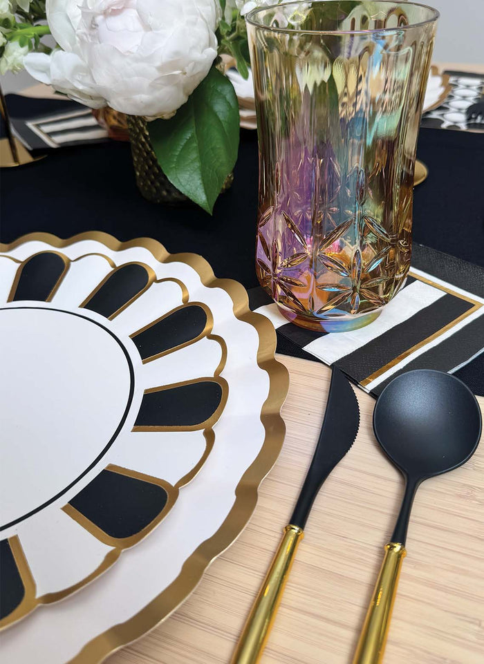 A close-up of an elegant table set with Sophistiplate Cordoba Dinner Plates (8pk), detailed with scalloped gold edges, paired with black and gold cutlery, a patterned glass, a monochrome napkin, and a white flower in a small vase.