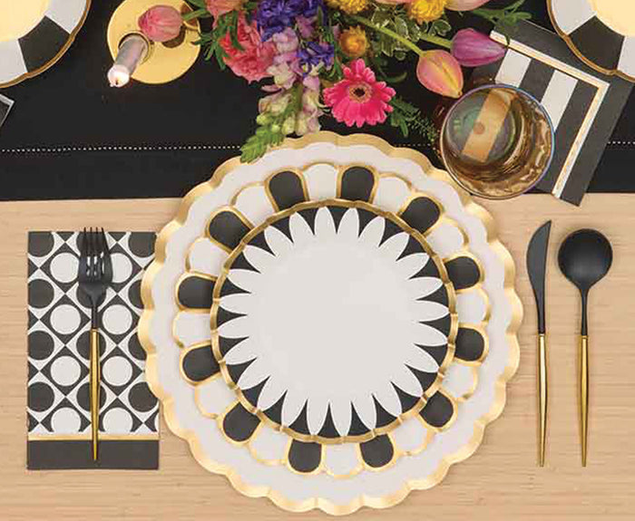A decorative place setting features Sophistiplate Cordoba Salad Plates/8pk, black flatware with gold handles, a patterned napkin, glassware, and a floral centerpiece on a light wooden table.