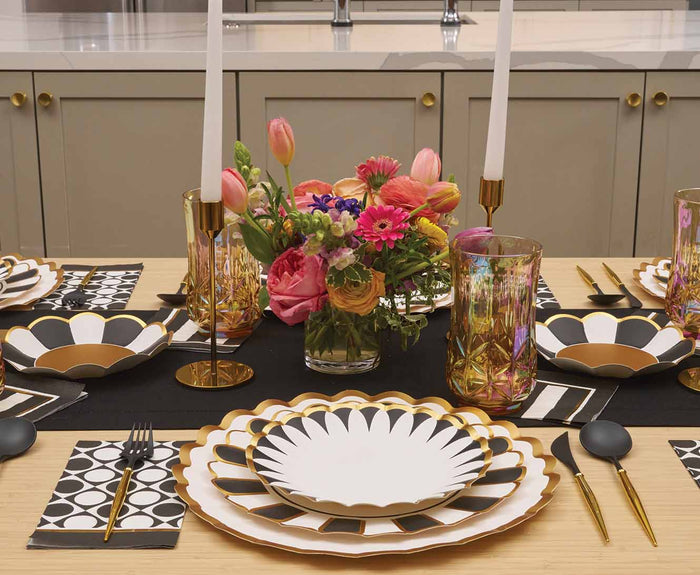 The Cordoba Salad Plates/8pk by Sophistiplate are set on a dining table with black cutlery, geometric napkins, gold candlesticks with white candles, a colorful floral centerpiece, and decorative glasses against a neutral background.
