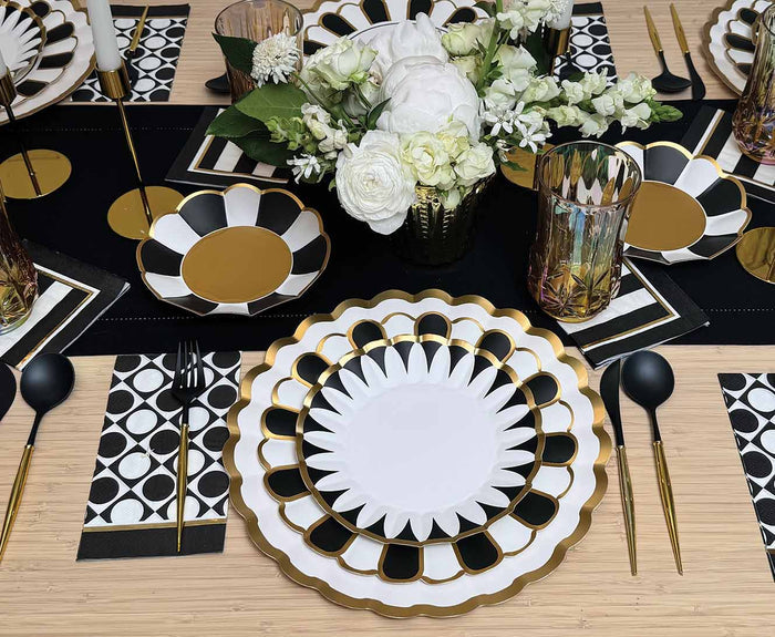 A modern table setting uses Sophistiplate Cordoba Salad Plates (8pk) with metallic gold trim plates, black cutlery, gold-accented glasses, patterned napkins, and a white floral centerpiece atop a black table runner.