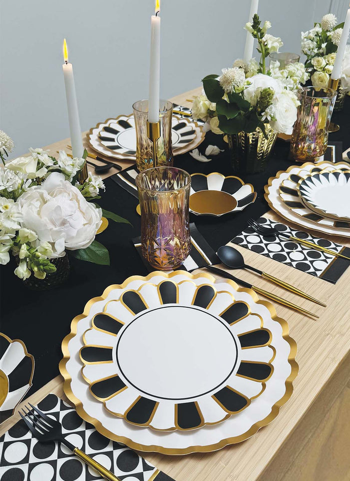 The Cordoba Table Setting from Sophistiplate LLC features gold and white designer disposable plates, black and white geometric napkins, gold flatware, purple glasses, white floral centerpieces, and tall white candles on a wooden table.