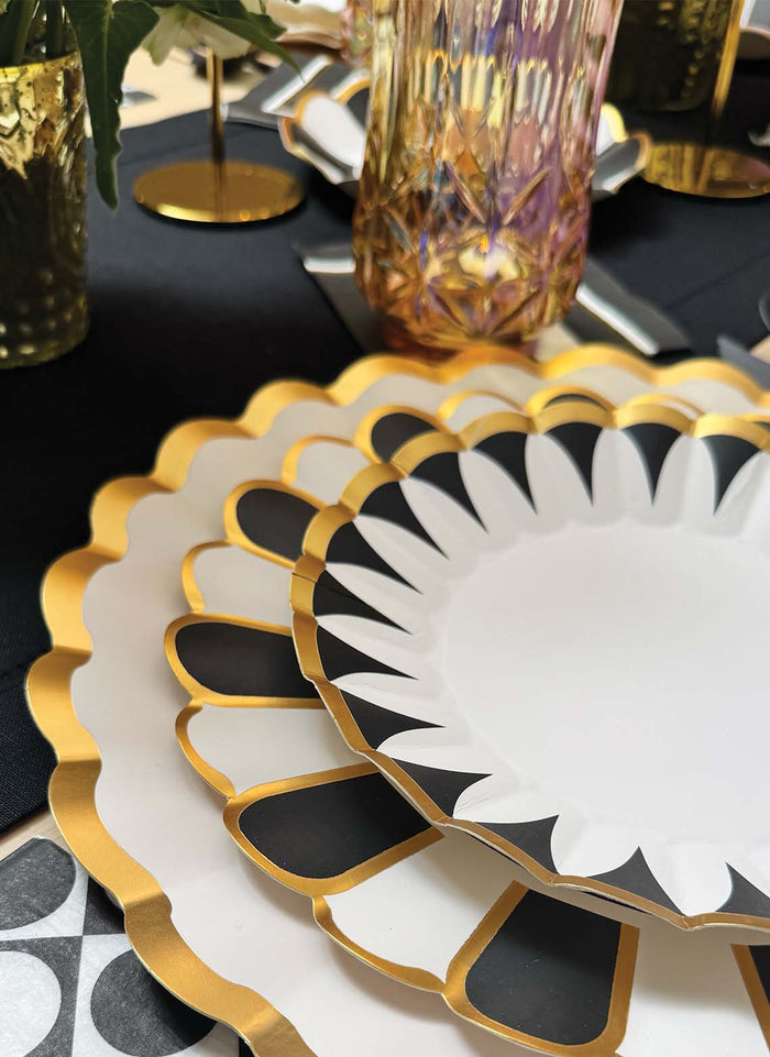 A close-up of the Cordoba Table Setting by Sophistiplate LLC, showcasing designer disposable plates with black, white, and gold scalloped designs, a gold-patterned tumbler, and gold accents on a black tablecloth.