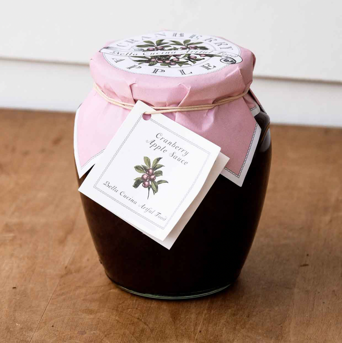 A glass jar of Bella Cucina Cranberry Apple Sauce, a sweet and tart Thanksgiving condiment, sits on wood with a pink paper cover, tied with string and a fruit-illustrated tag.