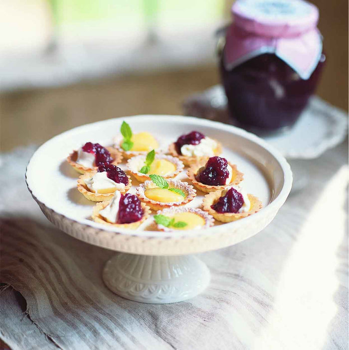 A white cake stand displays mini tarts garnished with cream, jam, and mint leaves. In the background, a jar of Bella Cucina Cranberry Apple Sauce—a classic Thanksgiving condiment—rests on a rustic surface.