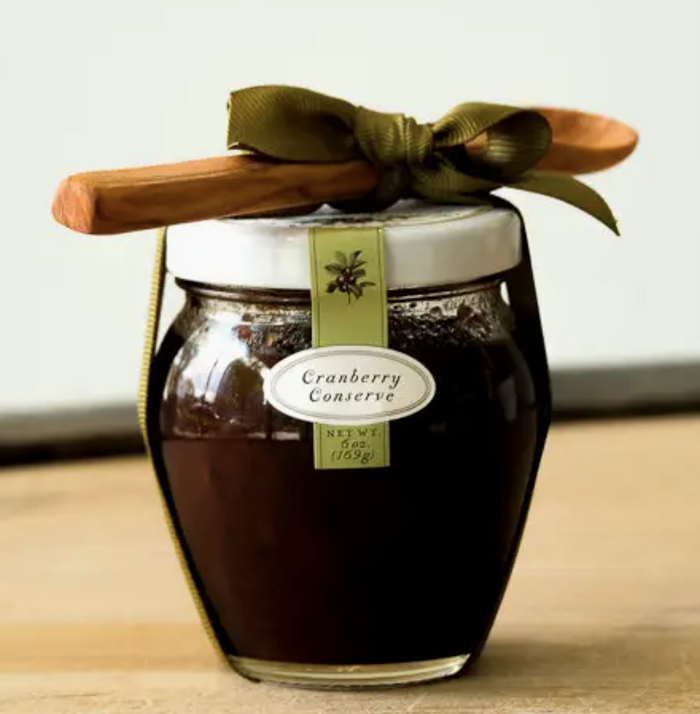 A glass jar of Bella Cucina Cranberry Conserve Gift w/ Spoon sits on a light wooden surface, adorned with a white lid, olive green ribbon, and a spoon—perfect as a sweet and tart Thanksgiving condiment.