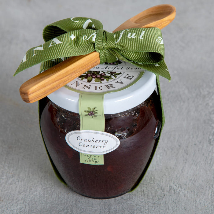 Bella Cucina’s Cranberry Conserve Gift w/ Spoon rests on a gray surface with a green ribbon and wooden spoon—an ideal sweet and tart sauce for your holiday table.