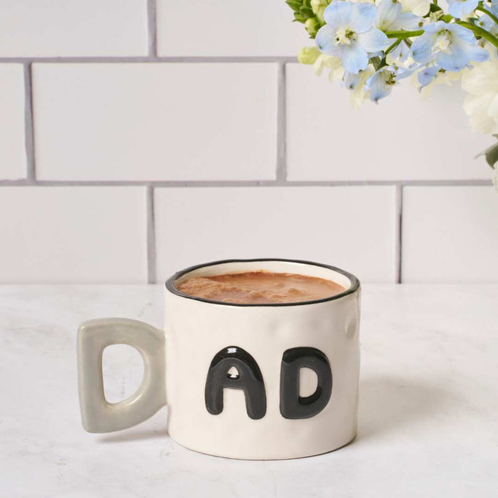 The Sophistiplate LLC Dad Mug + Hot Cocoa Gift Set sits on a white countertop filled with coffee, while soft blue and white flowers in the background create a cozy atmosphere, making this set an ideal gift for dads.