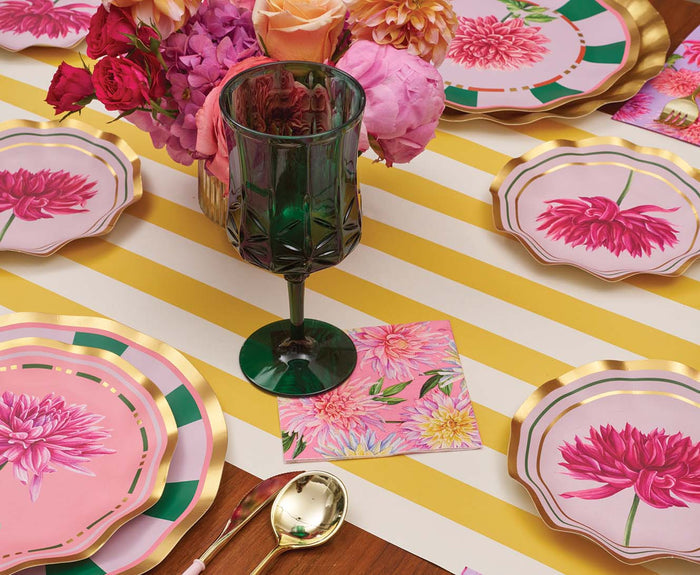 A table set with pink floral plates, a green glass, gold utensils, and a centerpiece of colorful flowers on a yellow-and-white striped tablecloth features the Sophistiplate Dahlia Garden Appetizer/Dessert Bowls (8pk), perfect for serving appetizers in style.
