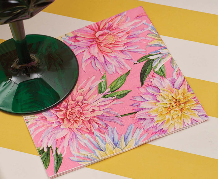 A green glass rests on a Sophistiplate Dahlia Garden Cocktail Napkin, featuring colorful dahlia flowers and green leaves, set atop a yellow and white striped surface.