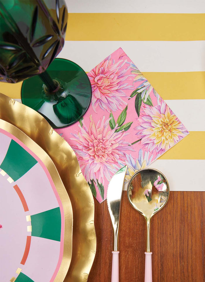 The Dahlia Garden Table Setting by Sophistiplate LLC features a green goblet, pink floral napkin, pink and gold cutlery, designer pink and gold plates, and a yellow-striped tablecloth on wood.