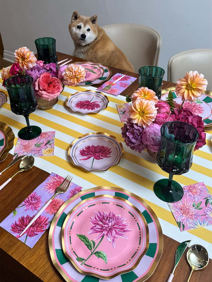 A Shiba Inu sits at a dining table set with pink floral plates, green glasses, and flower centerpieces. The table is adorned with a yellow and white striped runner, matching floral napkins, and Sophistiplate’s Dahlia Garden Appetizer/Dessert Bowls (8pk), perfect for serving appetizers or treats.