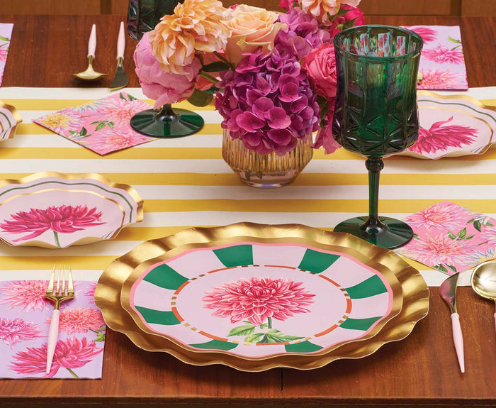 A table set for a meal showcases Sophistiplate Dahlia Garden Dinner Plates (8pk) with gold trim, green glasses, gold utensils, pink floral napkins, a yellow-striped runner, and vibrant floral arrangements.