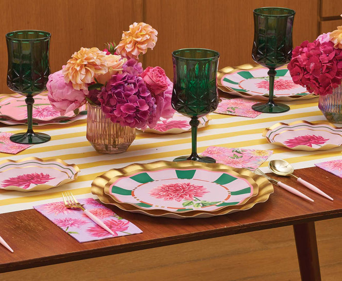 A wooden dining table set with Sophistiplate’s Dahlia Garden Dinner Plates (8pk), green glasses, pink utensils, pink floral napkins, and vases of pink and orange flowers on a yellow and white striped runner.