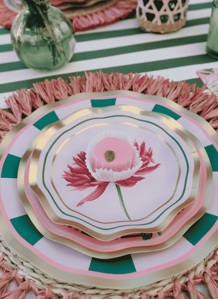 Layered Dahlia Garden Appetizer/Dessert Bowls from Sophistiplate, featuring pink, green, and gold-rimmed edges, are arranged on a woven placemat. The top bowl showcases a delicate pink flower design with a pink candy in a white wrapper at the center—perfect floral party bowls for your next gathering. Green vases decorate the background.