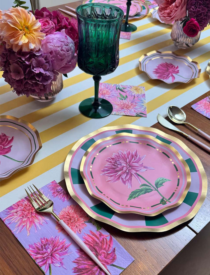 A colorful table setting with pink floral plates, gold utensils, green glassware, and a striped yellow-and-white table runner, decorated with vases of pink and purple flowers, is perfectly complemented by the Sophistiplate Dahlia Garden Appetizer/Dessert Bowls (8-pack) for serving appetizers.