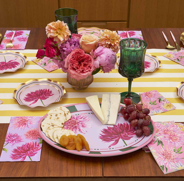 A colorful table is set with pink floral plates and napkins, a yellow-and-white striped runner, green glasses, a vase of pink and peach flowers, and the Sophistiplate Dahlia Garden Melamine Serving Platter with crackers, dried apricots, grapes, and cheese.