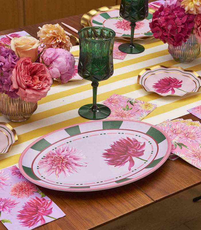 A wooden table features floral oval plates, green glasses, pink napkins, and vases of pink and purple blooms. A white and yellow striped runner and the Sophistiplate Dahlia Garden Melamine Serving Platter complete the setting.