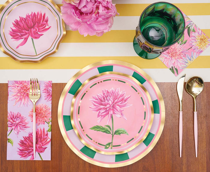A table set with floral pink plates, Sophistiplate Dahlia Garden Guest Towels, a green glass, gold and pink cutlery, and a pink flower centerpiece atop a white and yellow striped tablecloth.
