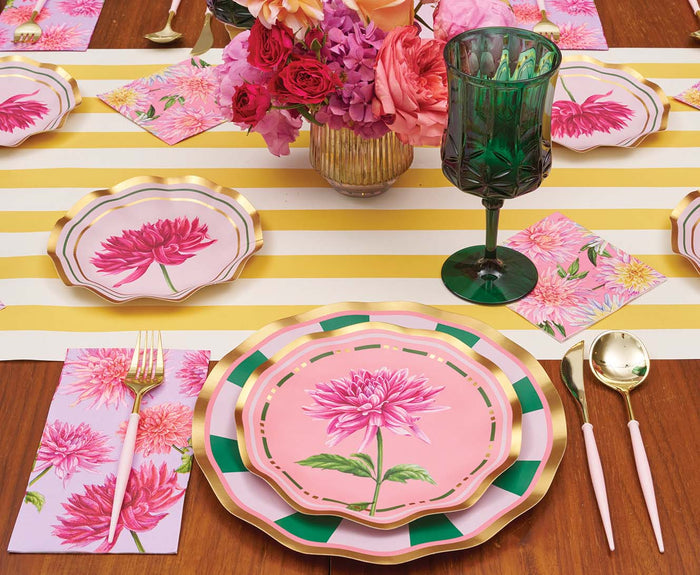 The Dahlia Garden Table Setting by Sophistiplate LLC displays designer floral plates, gold-trimmed utensils, green glassware, floral napkins, and a vase of blooms atop a yellow-and-white striped runner.