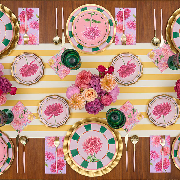 A table set for six features pink floral plates, gold utensils, green glasses, pink napkins, and a yellow-striped runner. Pink and purple flowers are elegantly arranged at the center, while Sophistiplate’s Dahlia Garden Appetizer/Dessert Bowls add a delightful touch to the tablescape.