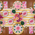 A table set for six features pink floral plates, gold utensils, green glasses, pink napkins, and a yellow-striped runner. Pink and purple flowers are elegantly arranged at the center, while Sophistiplate’s Dahlia Garden Appetizer/Dessert Bowls add a delightful touch to the tablescape.