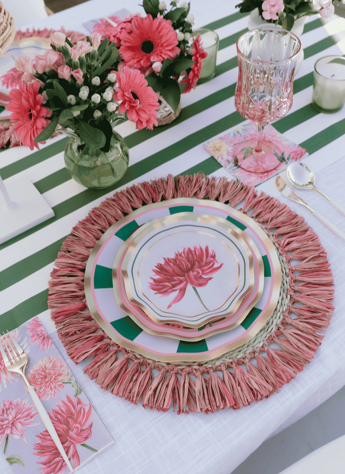 A table setting featuring pink floral plates, a pink fringed placemat, pink flowers in vases, a crystal glass, and a green-and-white striped tablecloth creates a delightful scene. Pink floral napkins and silverware are arranged neatly, while charming Sophistiplate Dahlia Garden Appetizer/Dessert Bowls (8pk) add extra elegance for serving appetizers.
