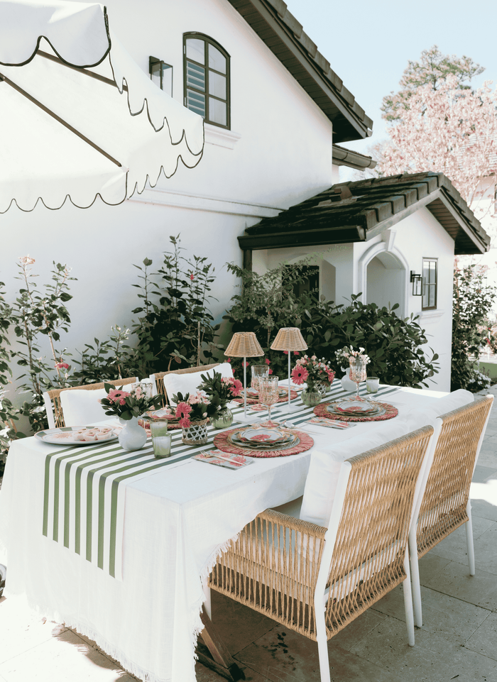The Dahlia Garden Table Setting by Sophistiplate LLC features a stylish outdoor table with pink blooms, a green and white striped runner, woven chairs, and designer disposable plates beneath a large umbrella beside a lush, white house.