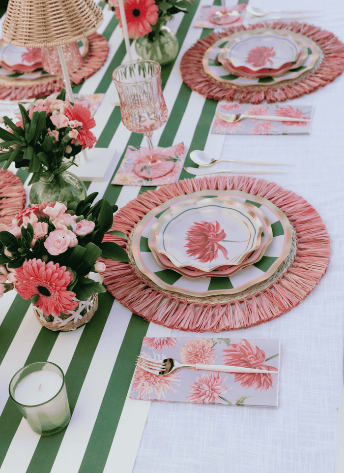 A spring-themed table is set with pink floral plates, napkins, and placemats, a green and white striped runner, pink flowers in vases, candles, and matching Sophistiplate Dahlia Garden Appetizer/Dessert Bowls (8pk)—perfect for serving appetizers—creating a fresh and elegant atmosphere.