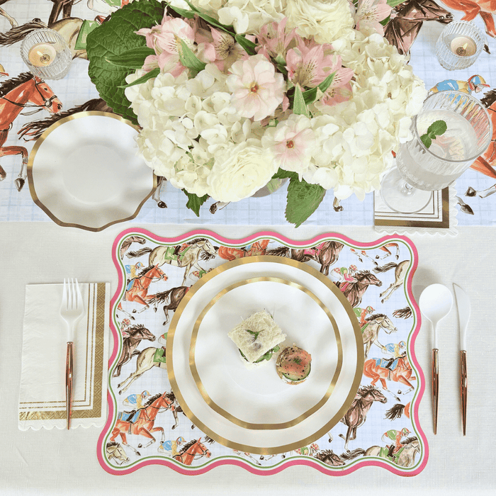 The Derby Table Setting by Sophistiplate LLC features decorative horse-patterned disposable plates, gold utensils, a white napkin with gold trim, a floral centerpiece, a water glass, and small sandwiches arranged on the main plate.