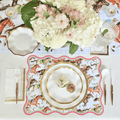 The Derby Table Setting by Sophistiplate LLC features decorative horse-patterned disposable plates, gold utensils, a white napkin with gold trim, a floral centerpiece, a water glass, and small sandwiches arranged on the main plate.