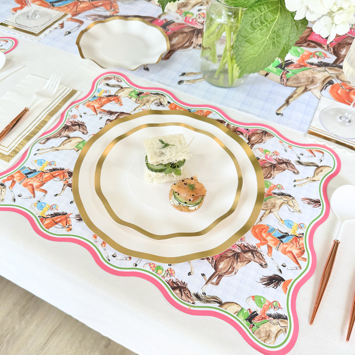 The Derby Table Setting by Sophistiplate LLC features equestrian-themed placemats, decorative disposable plates with a sandwich and appetizer, gold utensils, and a vase of green leaves and white flowers for a bright, elegant look.