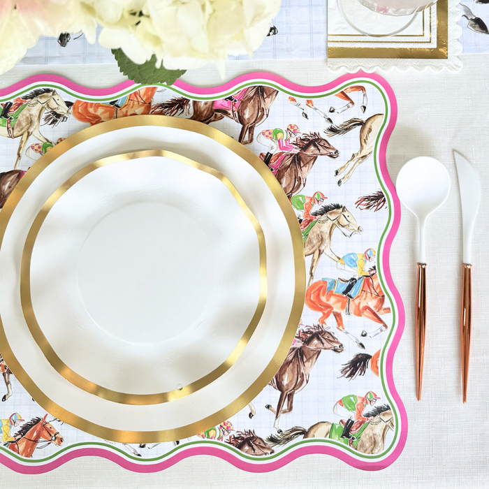 The Derby Table Setting by Sophistiplate LLC features decorative disposable plates with gold rims, a scalloped placemat with colorful horse and jockey designs, rose gold and white cutlery, a white napkin, and white flowers as an accent.