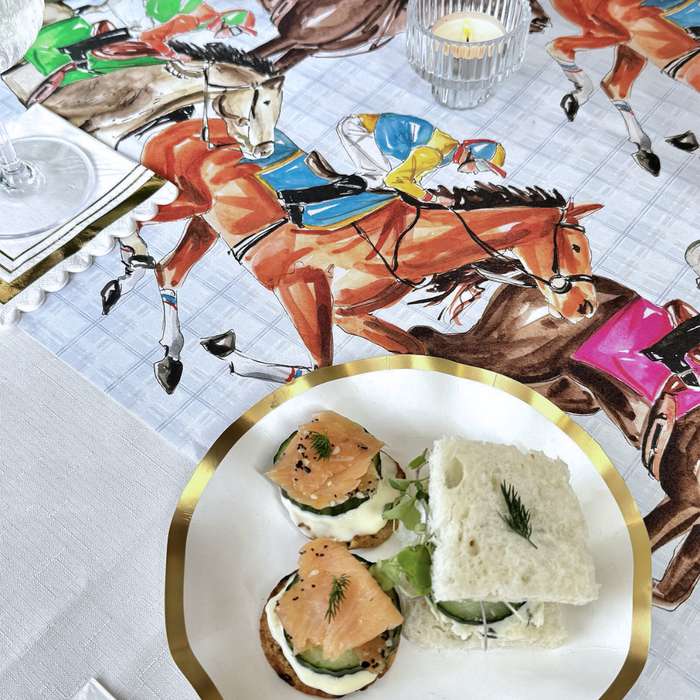 A plate with blinis and a sandwich rests on a table featuring the Rosanne Beck Derby Racehorses Table Runner (20x12") beside a lit candle in a glass holder.